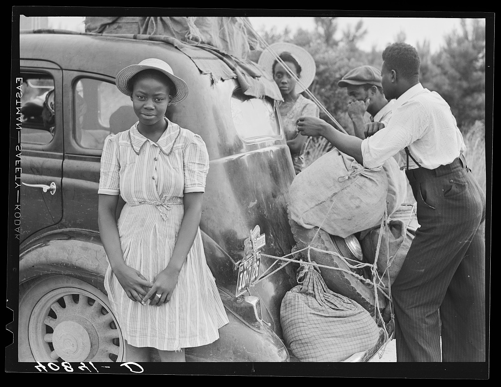 Black and white photograph of an African American family packing up their car during the Great Migration. A young woman is in front wearing a dress and smiling at the camera. Behind her, two men are putting luggage in the trunk of the car. In the background is a second young woman looking at the camera.