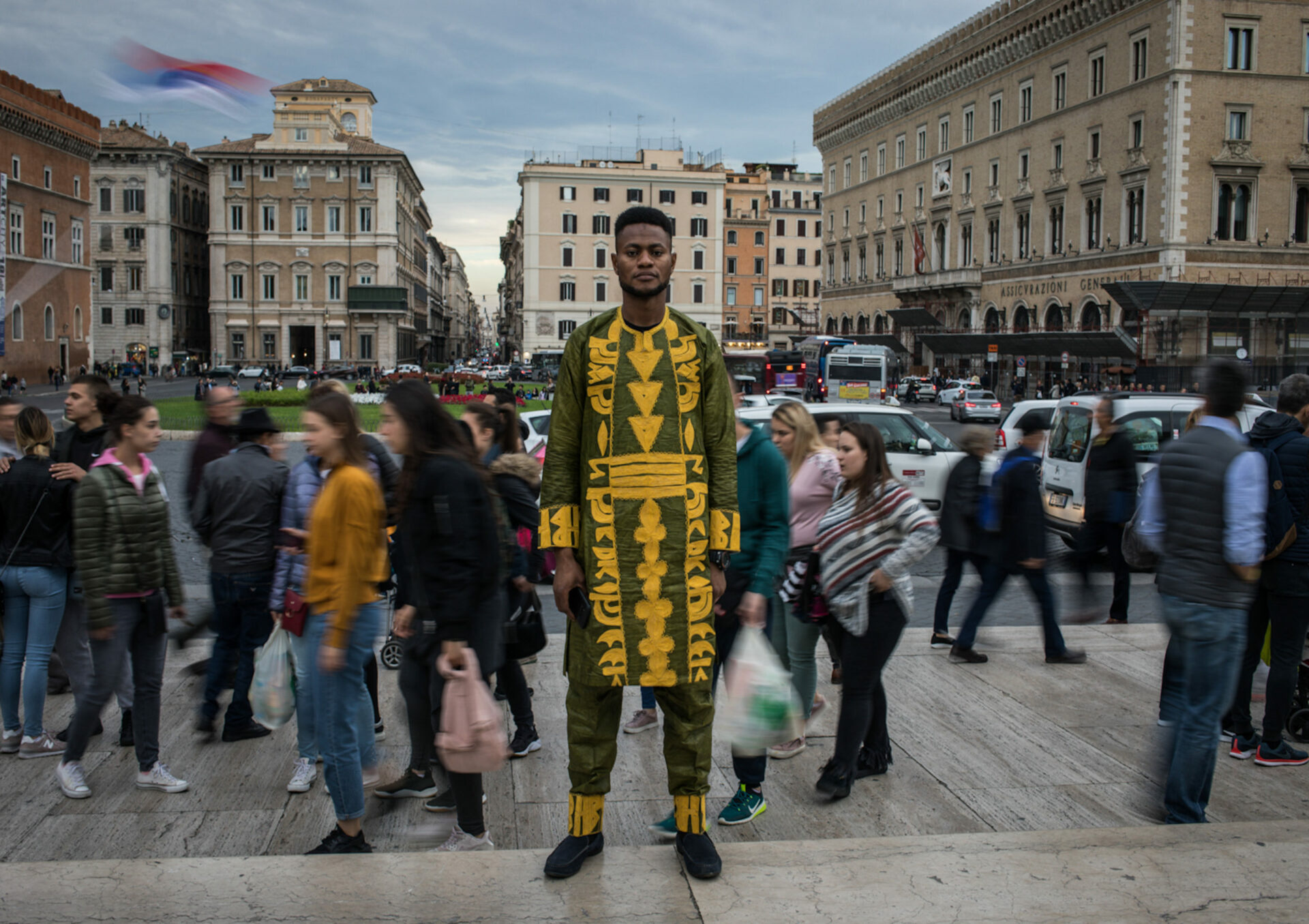 002_scaling-fences-african-migrants-rome Tall African man dressed in traditional gold and green dashiki stands in foreground on busy Roman pedestrian footpath