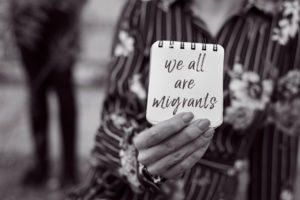 Closeup,Of,A,Young,Woman,Outdoors,Showing,A,Notepad,In closeup of a young woman outdoors showing a notepad in front of her face with the text we all are migrants written in it, in black and white