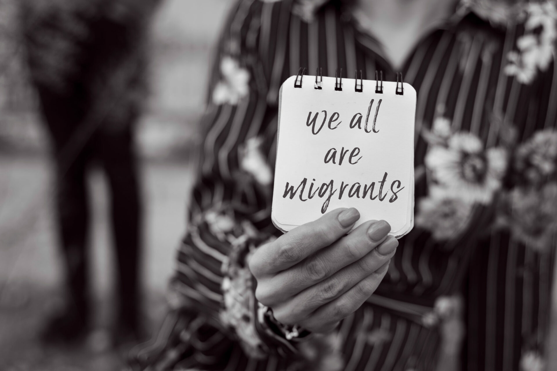 closeup of a young woman outdoors showing a notepad in front of her face with the text we all are migrants written in it, in black and white