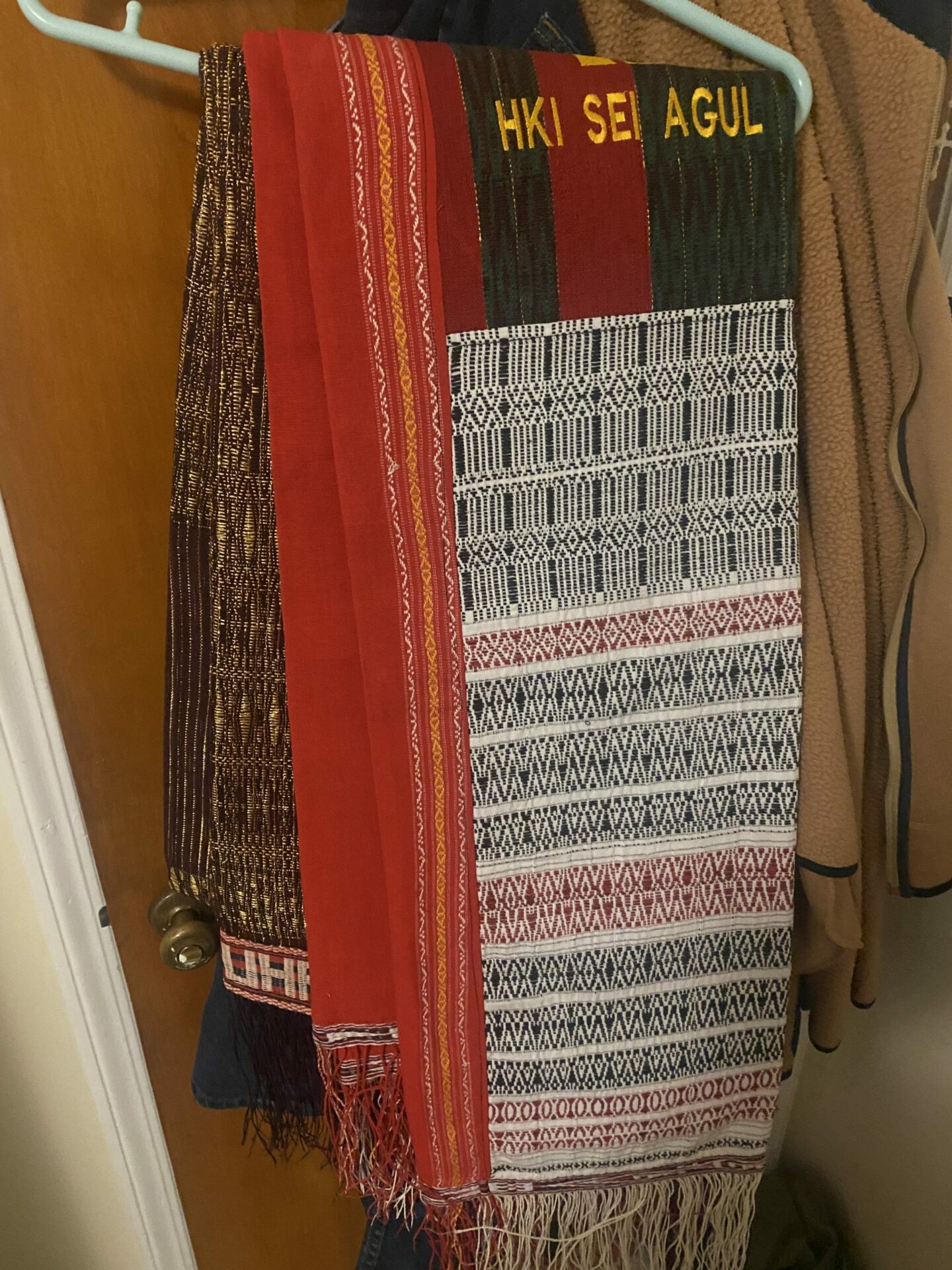 A photograph of an Ulos. The Ulos is a woven textile, with loose threads hanging out of the ends of both sides. It is red, white, and black