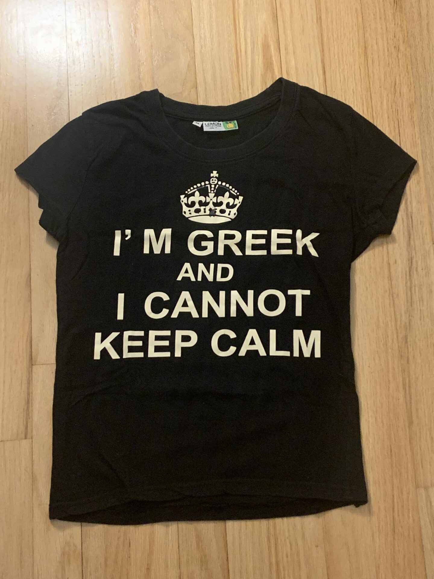A black shirt with a crown printed at the top and the words "I'M GREEK AND I CANNOT KEEP CALM" printed below it