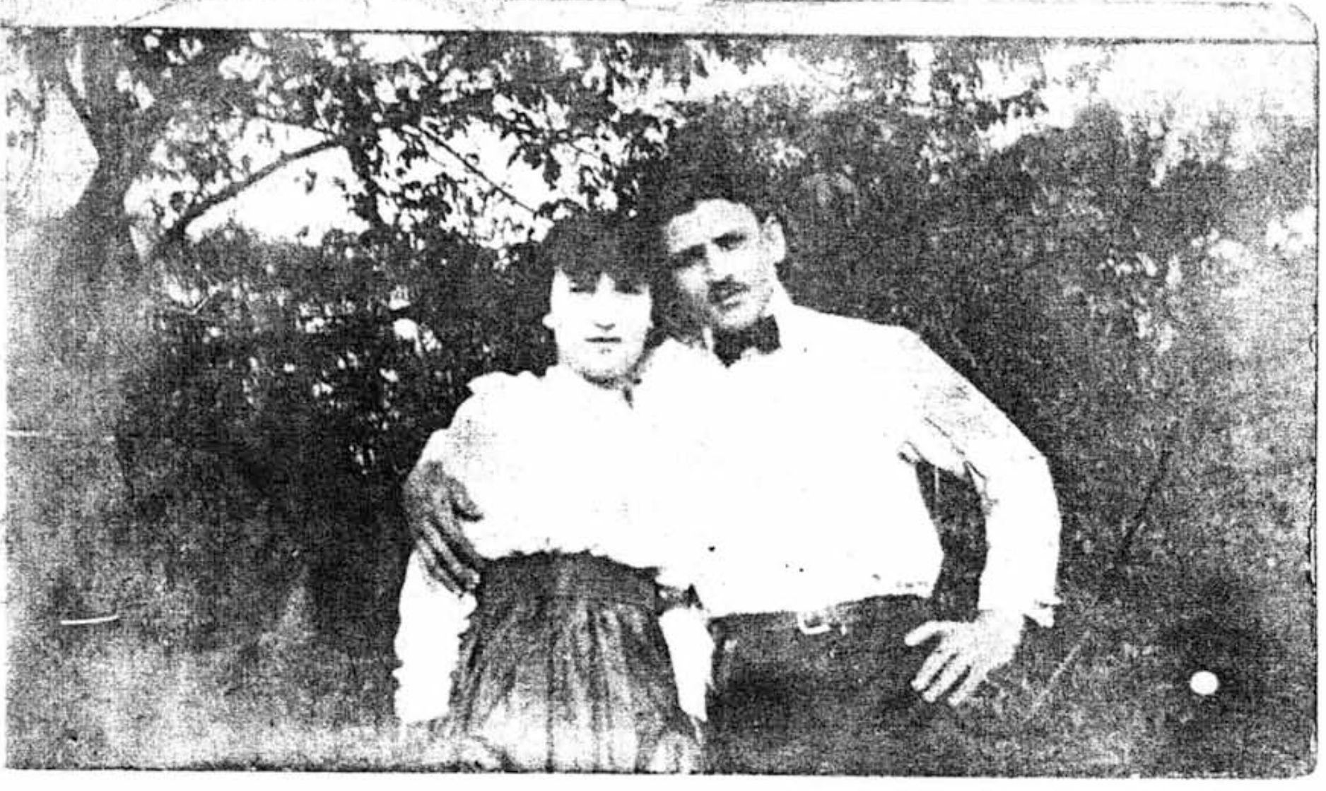 A young mustachioed dark-haired man in a black bowtie and white shirt, with his right arm around a young woman with white blouse and high-waisted skirt pose outdoors against a leafy background in this photo from 1917.